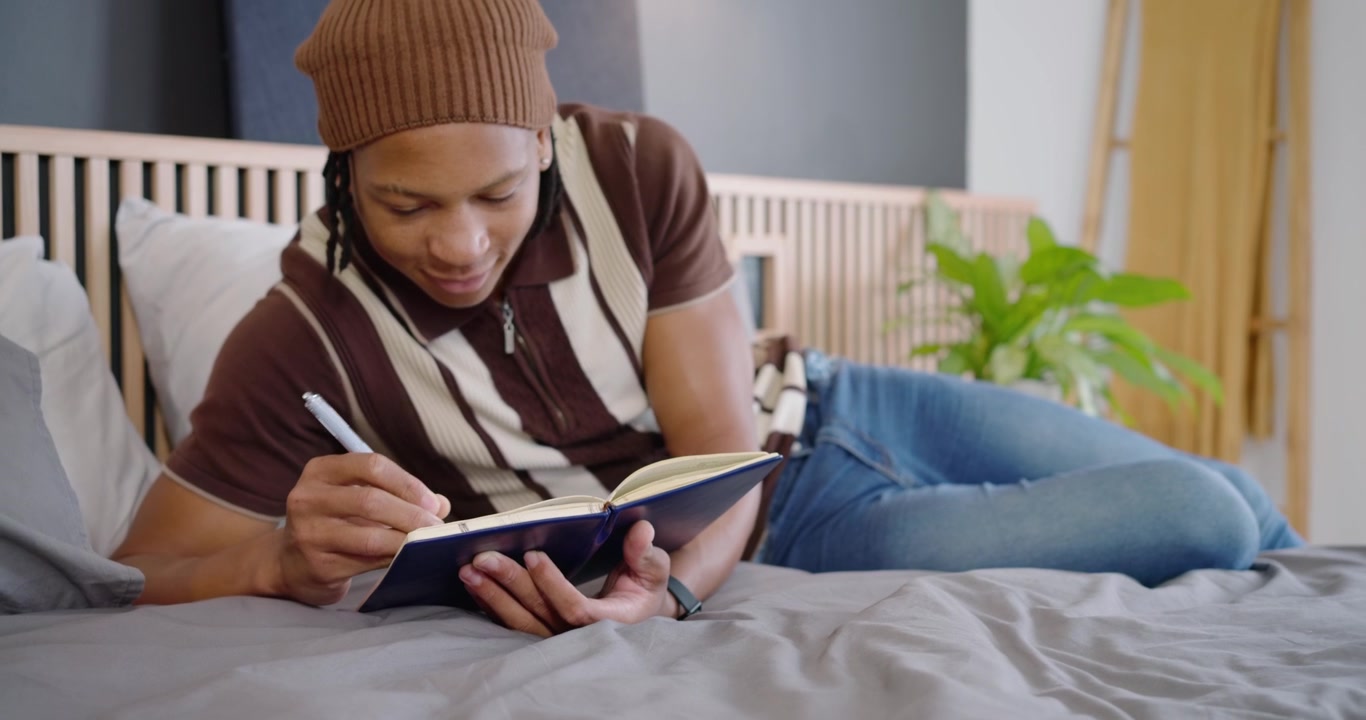 Uomo africano adulto che tiene un quaderno blu sul letto, che porta a scrivere con una penna, facendo una pausa e riflettendo.