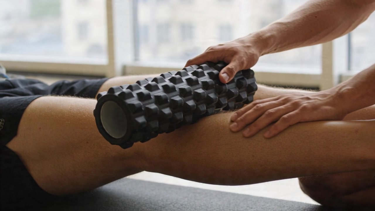 Istruttore che fa rotolare un rullo di schiuma nero lungo la coscia destra di un cliente in pantaloncini in palestra per terapia.