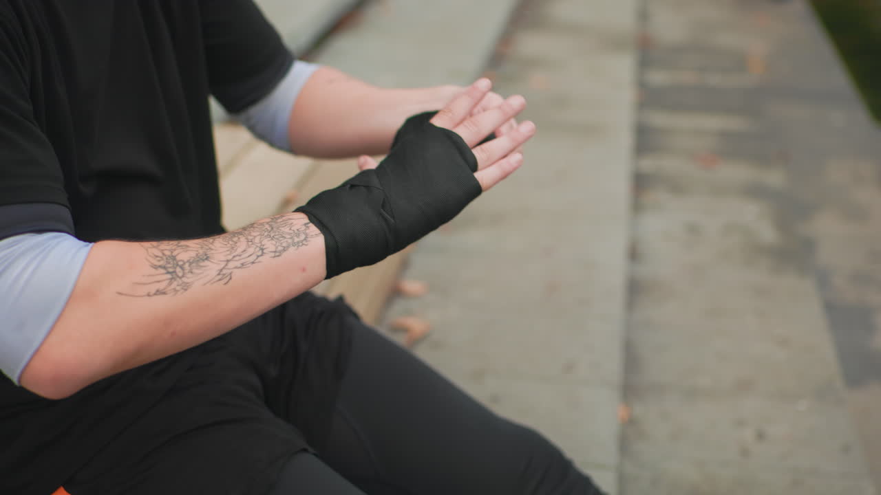 Kaukasischer Mann mit Tätowierungen, der sich auf Stufen eine Handbandage umwickelt, auf Betontreppen sitzend, schwarze Kleidung tragend, vorsichtige Bewegungen zur Sicherung der Handgelenkbandage, Nahaufnahme von Unterarmtätowierung und Knöcheln, düsteres urbanes Umfeld