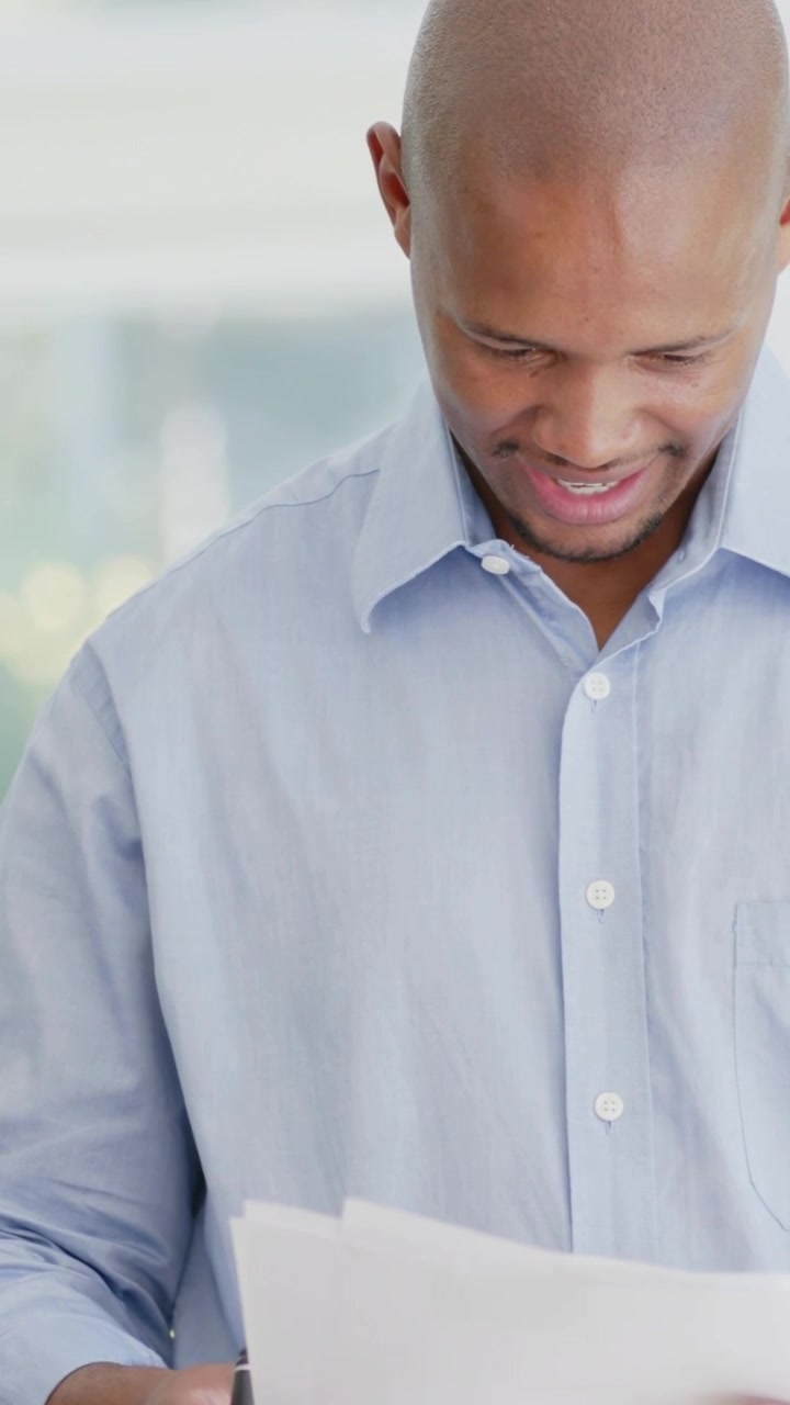 Vertical video: Picking up papers, man in light blue shirt reading, marking with pen by windows