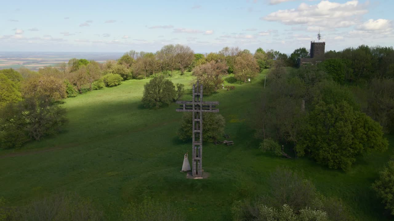 A large wooden cross monument stands prominently on a lush green hillside, surrounded by trees under a partly cloudy sky. In the background, a historic stone tower rises above the forest.