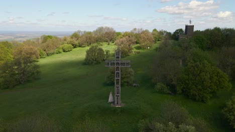 A large wooden cross monument stands prominently on a lush green hillside, surrounded by trees under a partly cloudy sky. In the background, a historic stone tower rises above the forest.