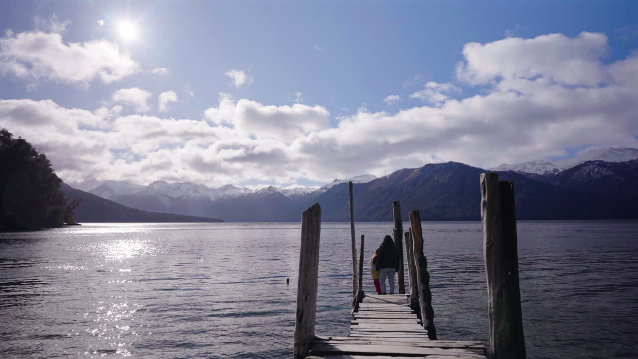 A mother and daughter stroll along a rustic wooden pier overlooking Lago Traful in Argentina