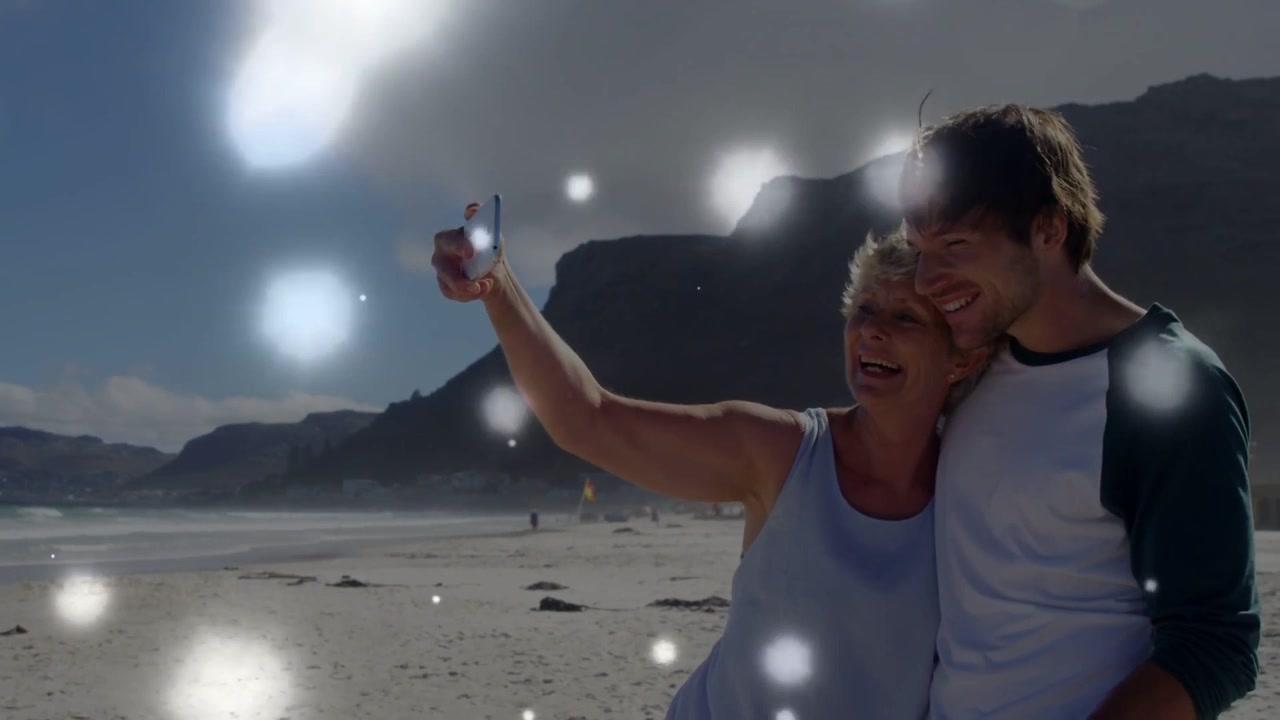 Mulher estendendo o telefone, homem envolvendo o braço para selfie enquanto bokeh flutua obscurecendo rostos para viagem.