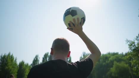 back view of a man holding a soccer ball up with his right hand, with a blurred view of people in the background