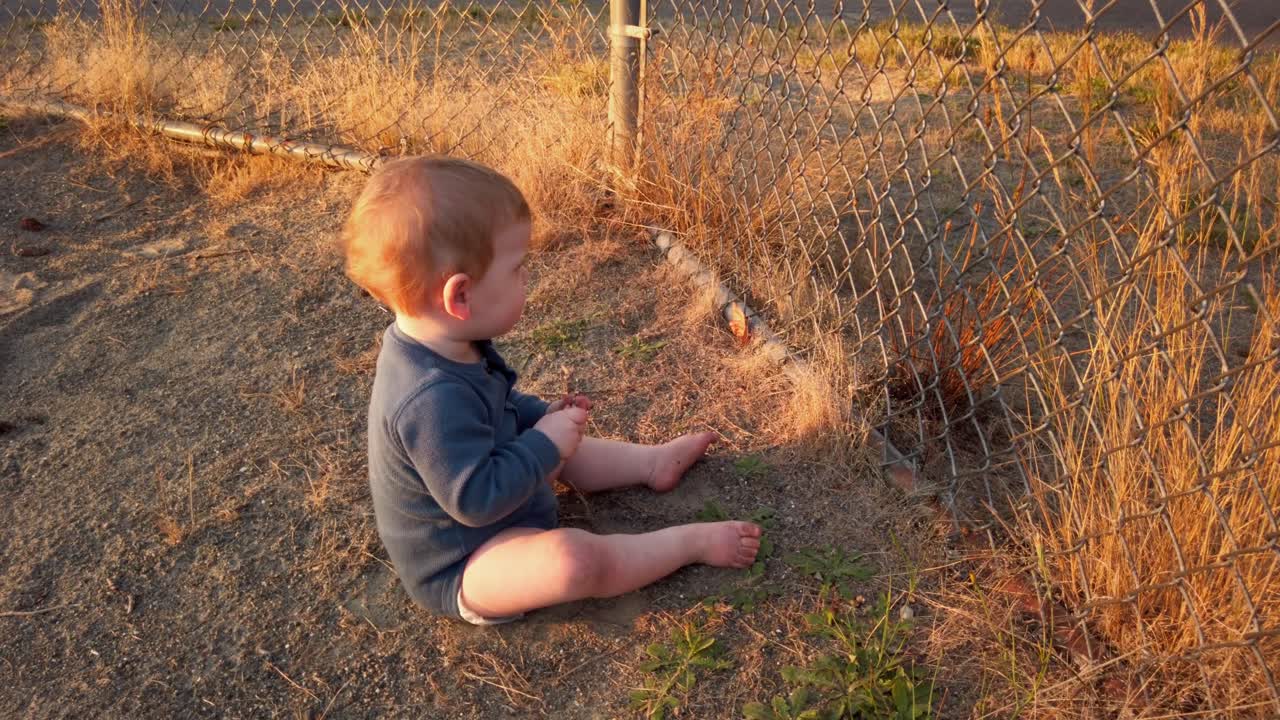 Happy toddler boy sits near home plate and plays with stick on fence