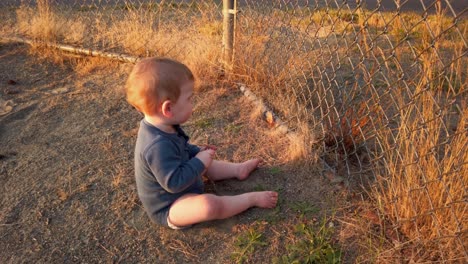 happy toddler boy sits near home plate and plays with stick on fence