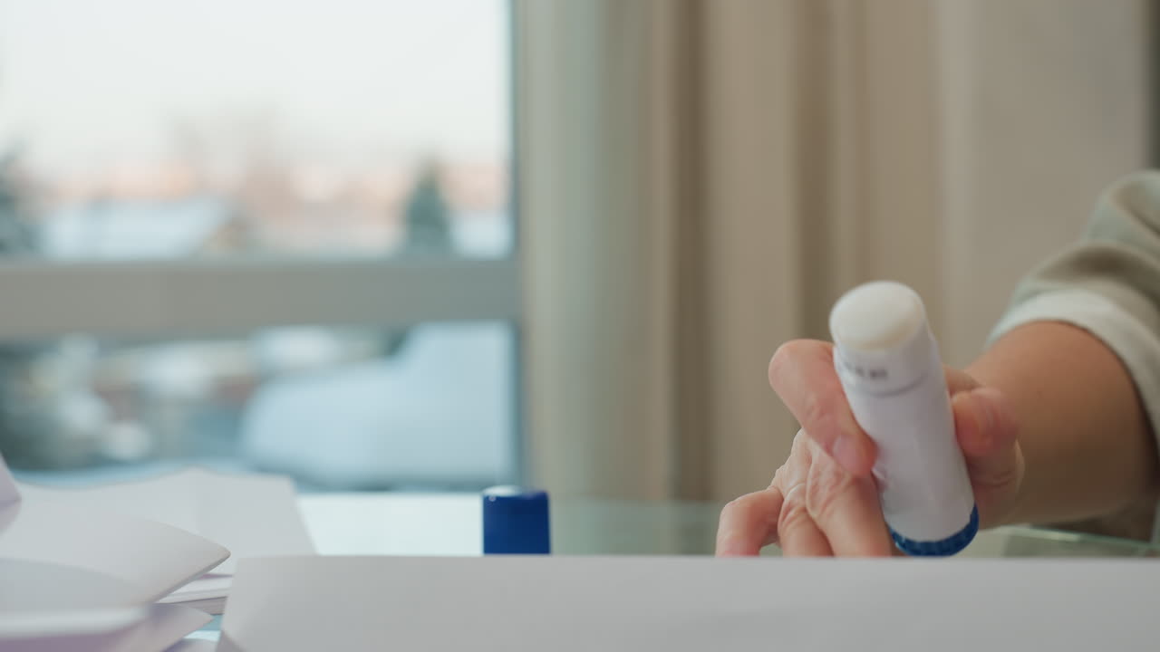 Hand view of older woman opening liquid glue container with white and blue cap and applying it on paper, close-up of hands engaged in crafting activity with focus on glue application
