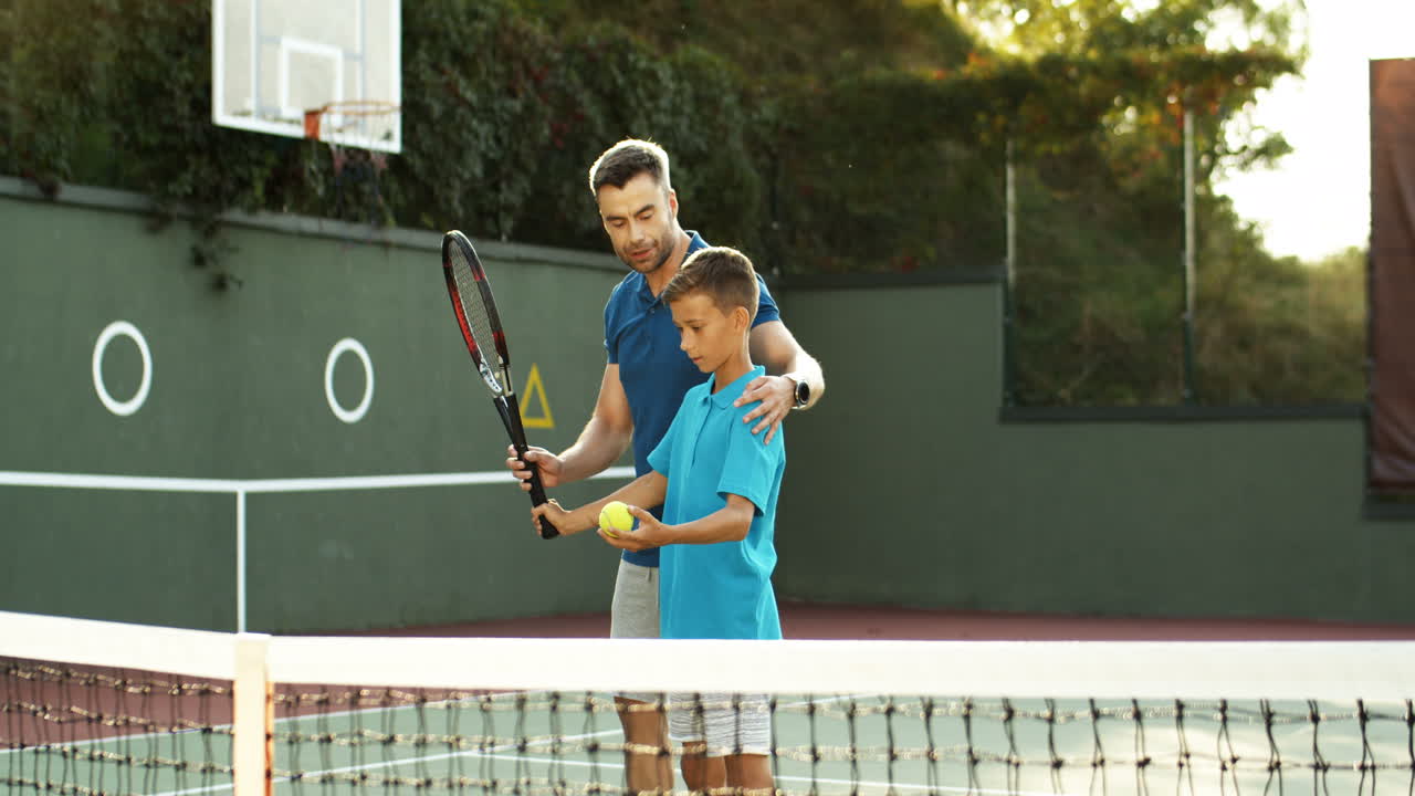 Man Teaching His Teen Son How To Hit Ball With Racket On A Tennis Court On A Summer Day