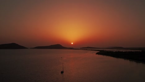 aerial reverse dolly above anchored sailboat as ball of sun sets casting warm gradient red rays into sky above pylos greece