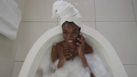 Mujer-Atractiva-Afroamericana-Sonriente-Relajándose-En-El-Baño-Y-Hablando-Por-Teléfono-Inteligente-En-El-Baño