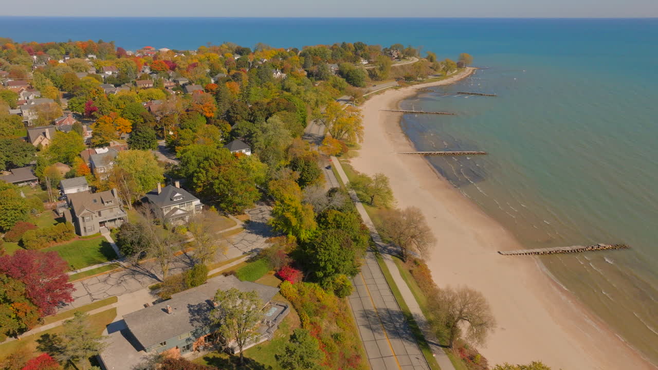 Drone aerial with pan and drift left over Lake Michigan beach toward Sheboygan, Wisconsin neighborhood homes and colorful autumn trees at peak foliage on a clear, pretty day