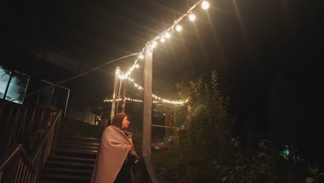 woman in blanket leans on stairs railing. lonely woman looks at distance during walk at nights in fresh air. observing nocturnal wildlife during rest on camping site