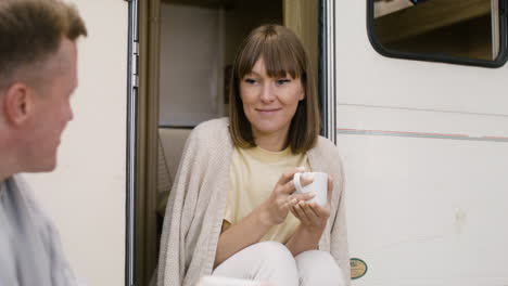 happy woman sitting on the stairs of campervan, holding coffee mug and talking with man