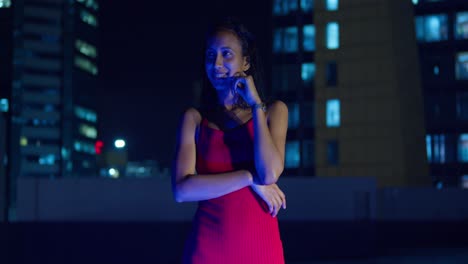 a young girl, dressed in a red dress, is on a rooftop at night with city buildings in the background