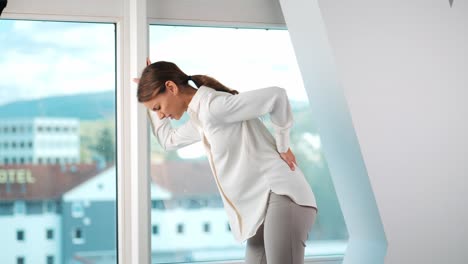 young woman feels back pain in front of window in modern apartment office