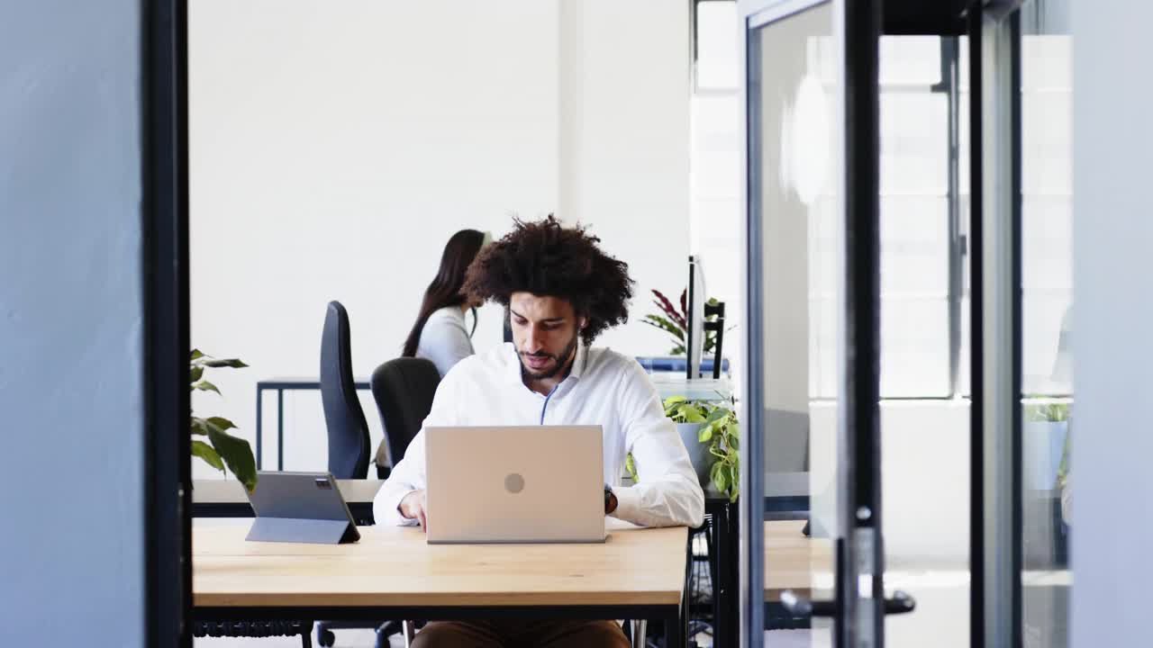 Man typing on laptop, pausing, responding to green code, adjusting headset, coworker behind in tech