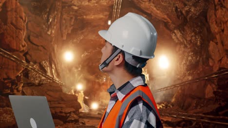 close up side view of asian male engineer with safety helmet working on a laptop and looking around while standing in underground mine tunnel
