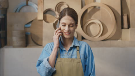 Portrait-of-caucasian-woman-in-apron-talking-on-the-phone-in-carpentry-workshop