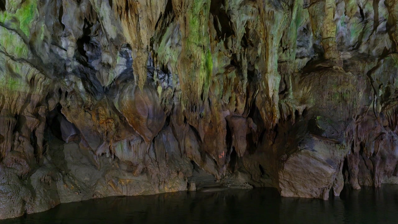Underground cave river flows through limestone chamber with hanging stalactites and mineral-stained rock formations. Static wide angle shot captures the natural geological features and dark water.