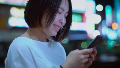 portrait of the attractive japanese girl with piercing and wearing casual clothes uses smartphone. in the background big city advertising billboards lights glow in the night.