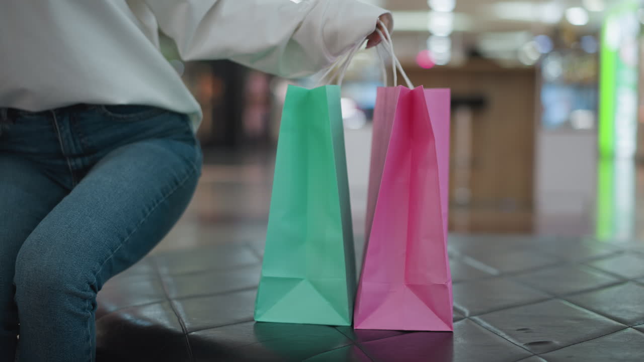 bolsas de compras rosas y menta colocadas en una superficie de baldosas, ligeramente inclinadas, con un fondo borroso del centro comercial, una mujer en vaqueros se sienta cerca en el asiento de cuero