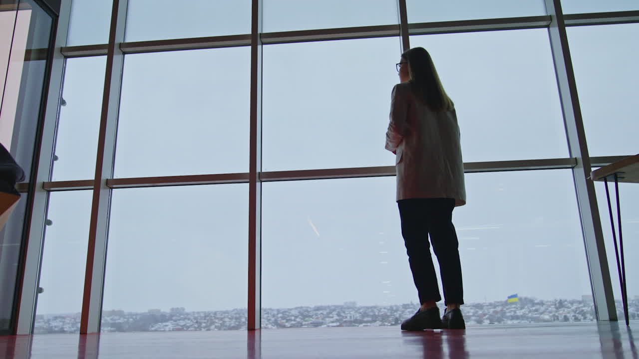 Long-haired lady in suit walks near the panoramic windows. Young woman looks at the city panorama. Low angle view.