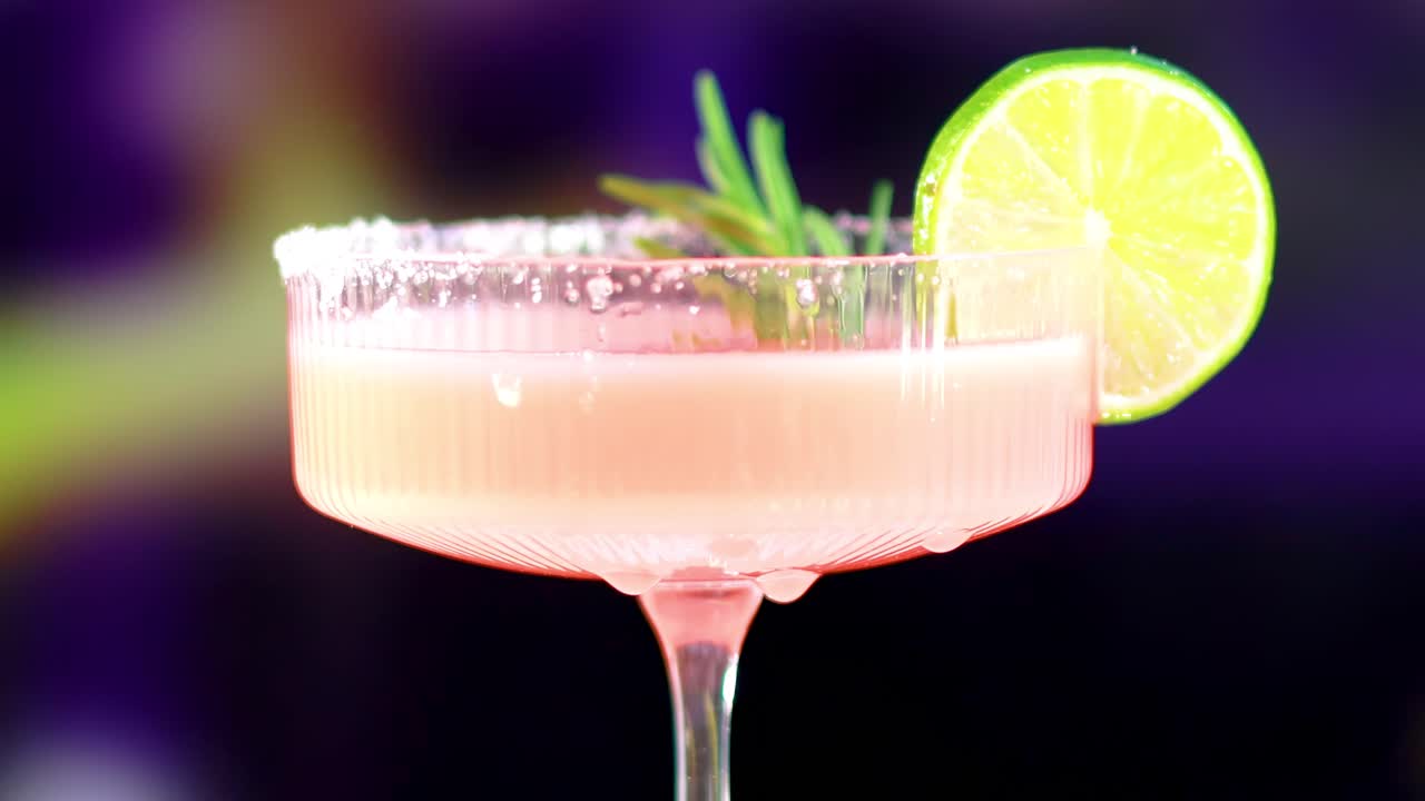 A detailed view of a pink cocktail garnished with lime and rosemary against a vibrant background.