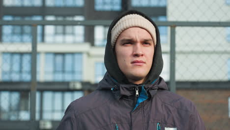 white man in hoodie and beanie holding ball in right hand walks through bar fence with sunlight reflecting on him, tall office building in background