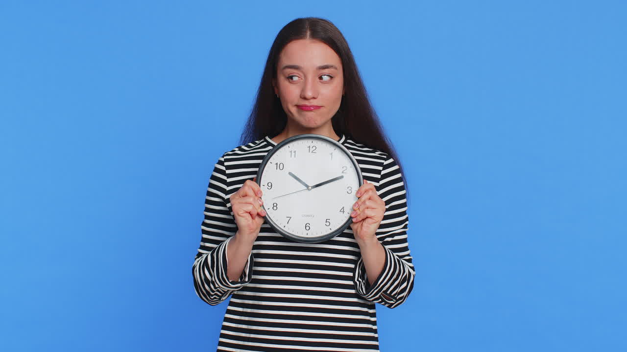 Serious strict young woman holding office clock hiding behind checking time on watch obscuring face