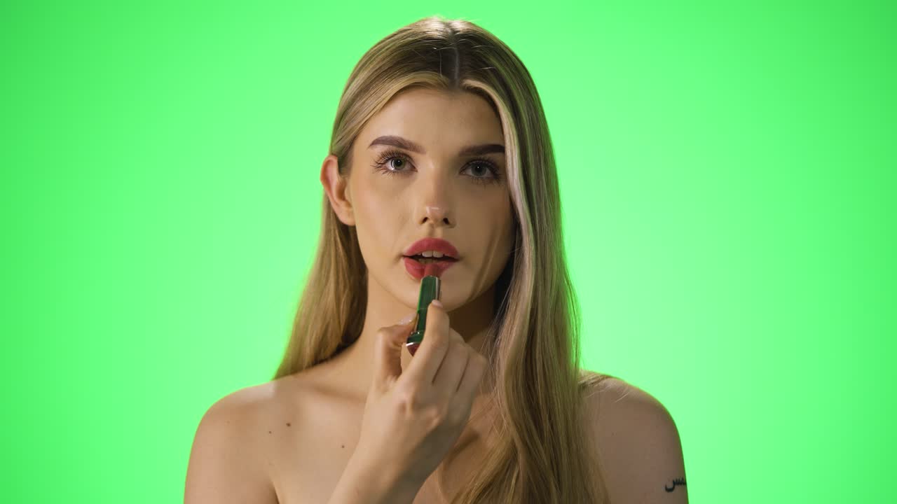 disparo estático de una joven bonita mujer aplicando su lápiz labial rojo mientras se prepara para la noche y luego dando un beso aéreo a la cámara con su mano frente al fondo verde en cámara lenta