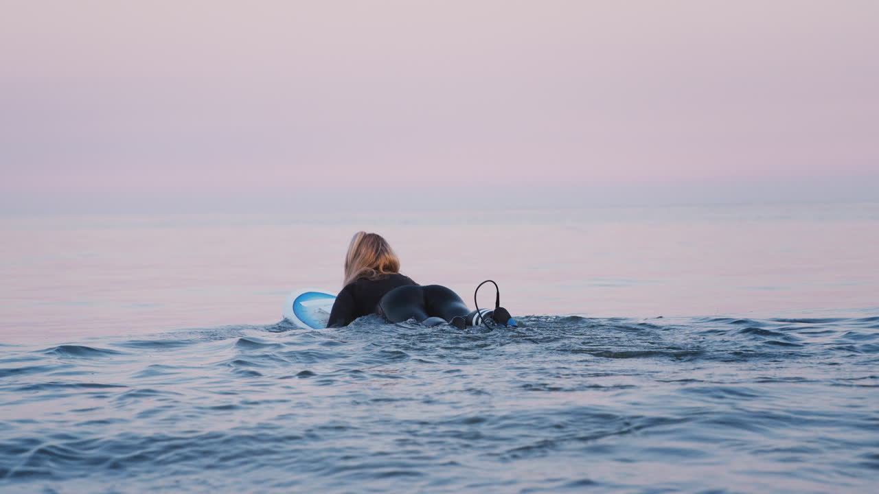 Rear View Of Woman Wearing Wetsuit Paddling Surfboard Out To Sea