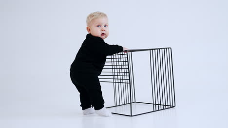 little blond child in black suit stands holding by the grate. kid turns to camera looking surprised. white backdrop.
