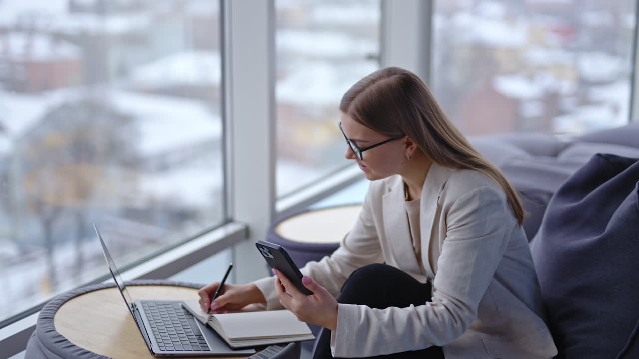 Female holding a phone in front of her and speaking to someone. Pretty lady taking notes in her book during conversation. Windows in blur at the backdrop.