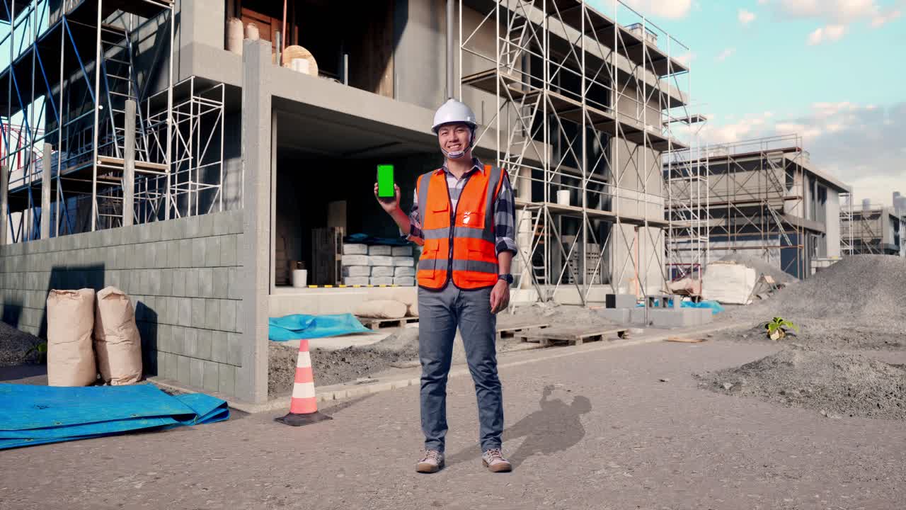Full Body Of Asian Male Engineer With Safety Helmet Smiling And Showing Green Screen Smartphone To The Camera While Standing At Building & Construction Site in Progress to New House
