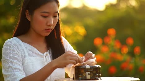 a serene moment: a young woman carefully unwrapping a delicate gift amidst a beautiful outdoor setting at sunset, surrounded by lush greenery and vibrant flowers