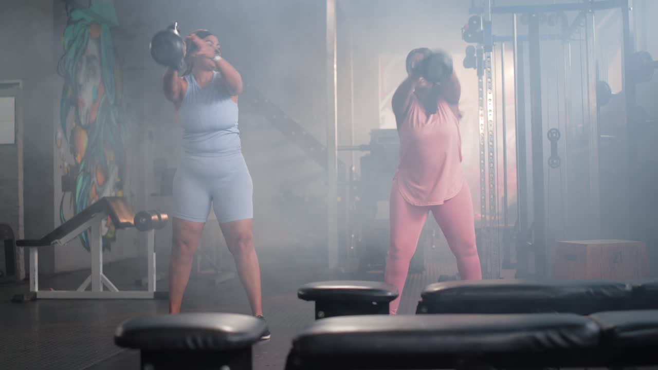 Two midadult women in activewear picking kettlebells swinging and pressing overhead in gym syncing