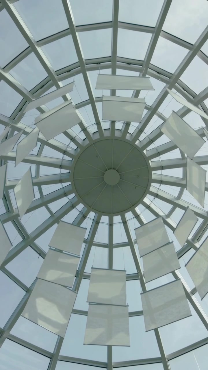 Video vertical: Paneles suspendidos balanceándose y girando bajo un tragaluz de cúpula de cristal en un atrio por la brisa.