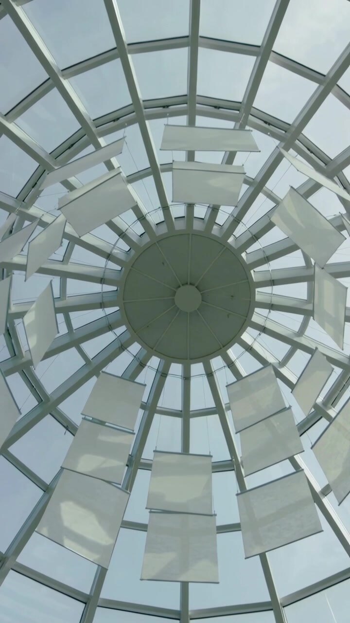 Video verticale: Pannelli sospesi che ondeggiano e ruotano sotto un lucernario a cupola di vetro nell'atrio a causa della brezza.