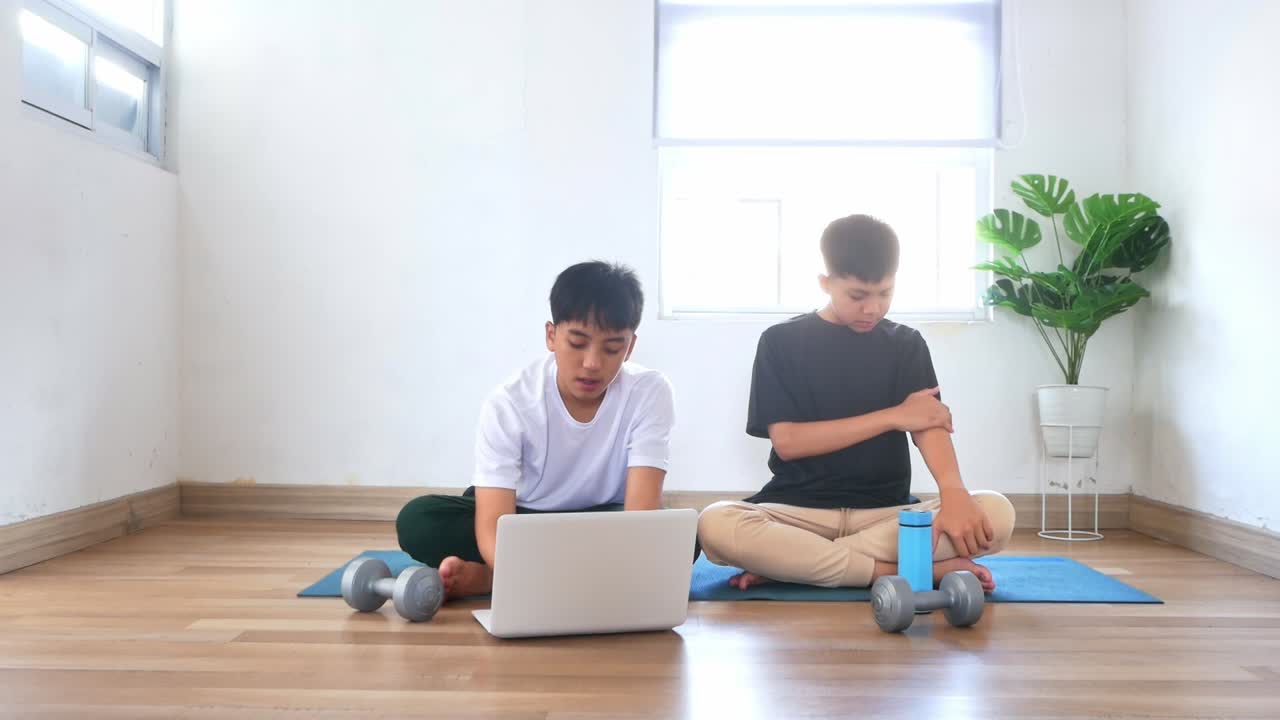 リビングで運動する前に、オンラインのワークアウト動画をノートパソコンで選んでいる若いアジア人の兄弟