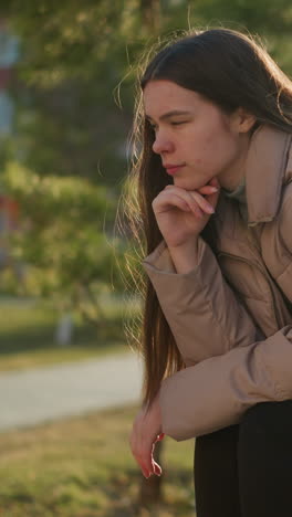 una joven con una chaqueta de melocotón, camisa interior gris y pantalones negros se sienta en un banco del parque, descansando su barbilla en una mano en profundo pensamiento. sus patines están colocados a su lado