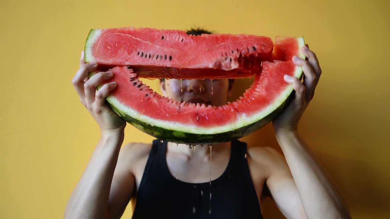Uomo in canottiera nera che alza un'anguria per allinearla alla bocca davanti a una parete gialla, con il succo che cola.
