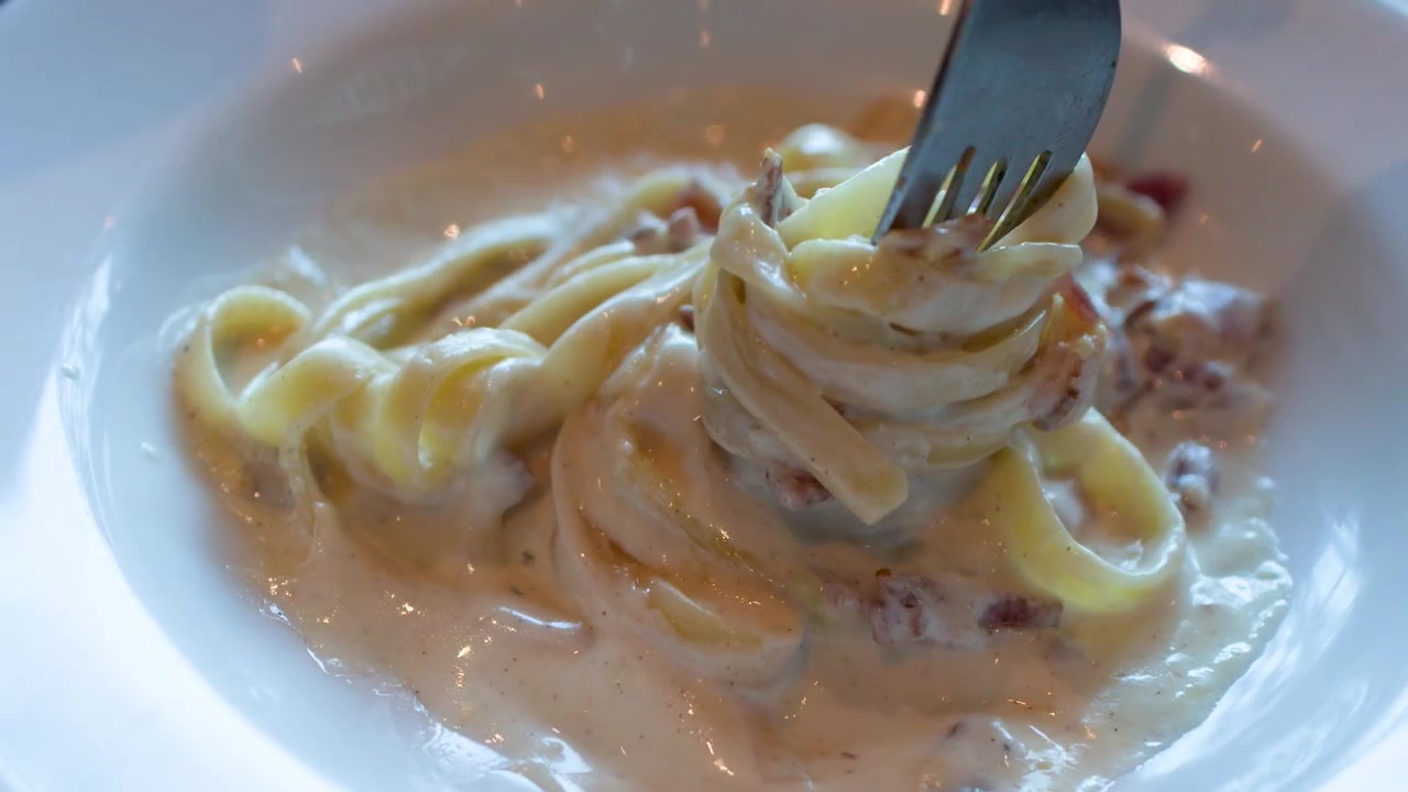 A fork twirls creamy fettuccine carbonara with bacon bits in a white ceramic bowl