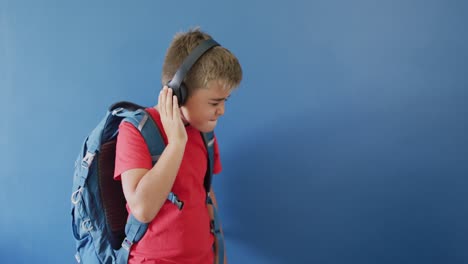 caucasian boy wearing backpack and using headphones on blue background, copy space, slow motion