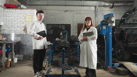 two inventors wearing lab coats and protective eyewear stand near an engine in mechanical workshop, holding tablets for data recording, bright backlight highlights industrial workspace