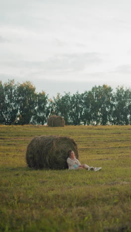 vista distante de la dama sentada en el suelo, descansando contra una gran bala de heno en tierras de cultivo abiertas, paisaje rural pacífico con balas de heno esparcidas y luz cálida de la tarde