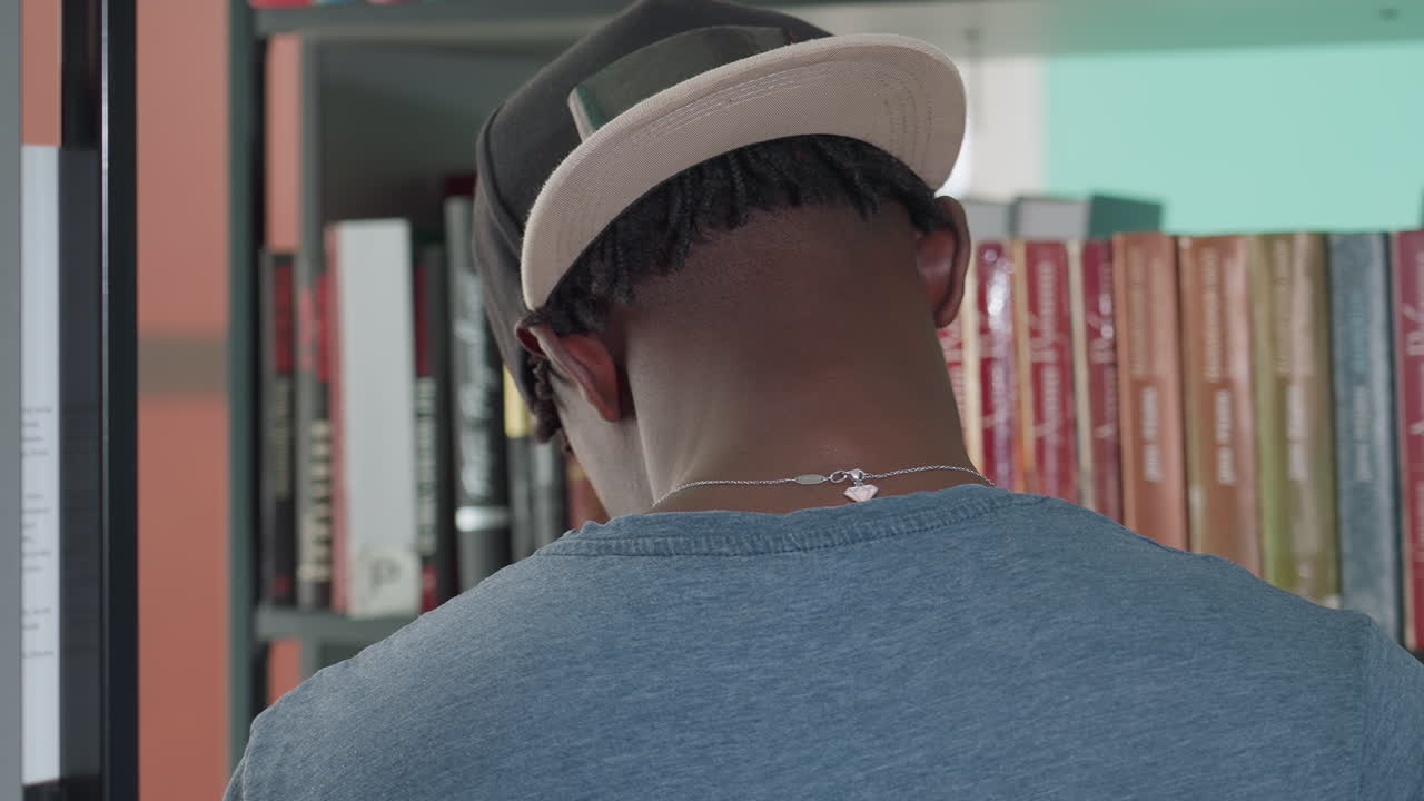 Back view of man in casual clothing and backward cap standing in library, facing bookshelves filled with colorful spines, appearing focused and engaged in quiet study