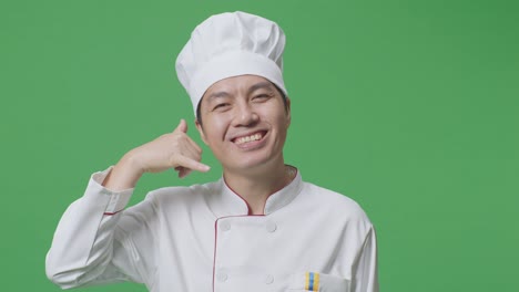 Close Up Of Asian Man Chef Smiling And Making Call Me Gesture To Camera While Standing In The Green Screen Background Studio
