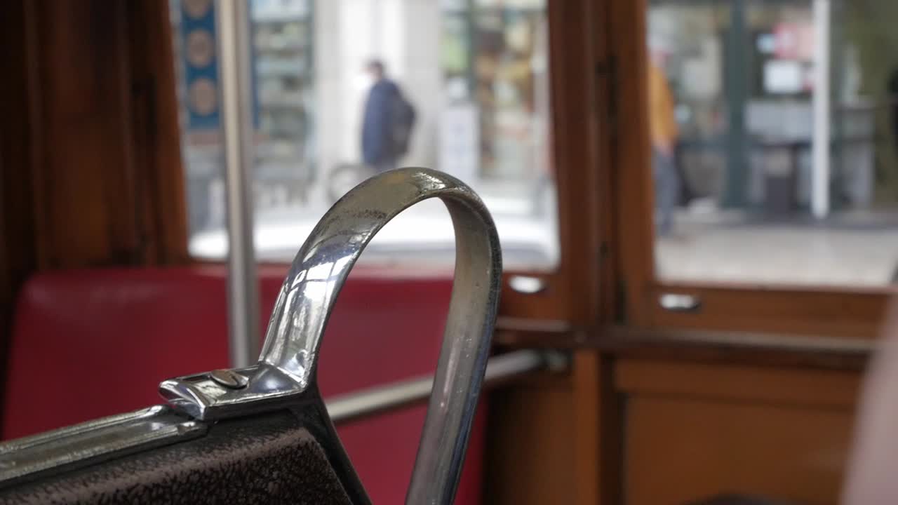 View of Lisbon streets from a tram in transit during a commute. Seat's handle focused on frame and slightly unfocused view of wooden interior and streets through the window. Cars, shops and people.