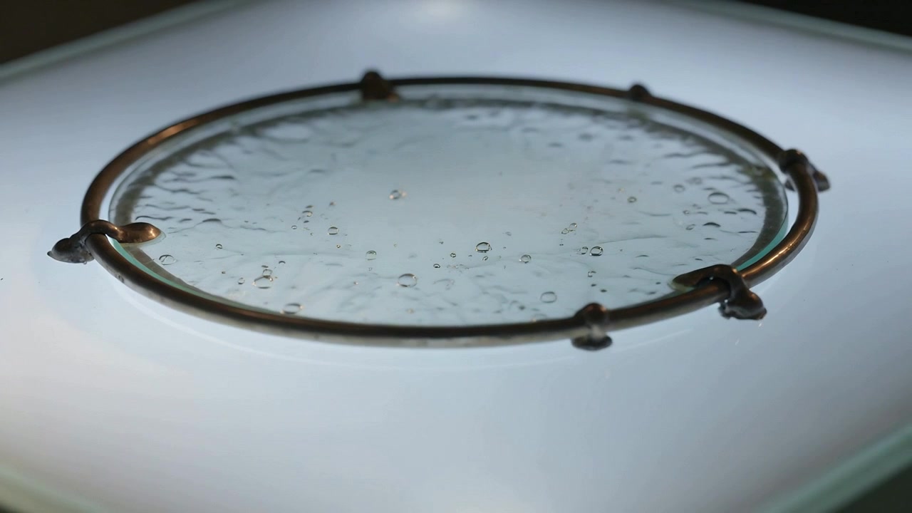 Sich bewegende Kamera enthüllt Tröpfchen auf einer runden Glasscheibe auf einem Leuchttisch und hebt dabei Metallringklemmen hervor.